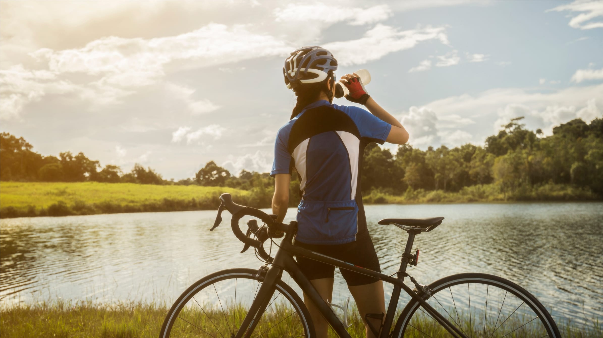 A woman cyclist taking a break next to a lake. She has a high end racing bike leaning against her whilst she takes a drink of Seltza water from a sports bottle. In the background are trees and grass.