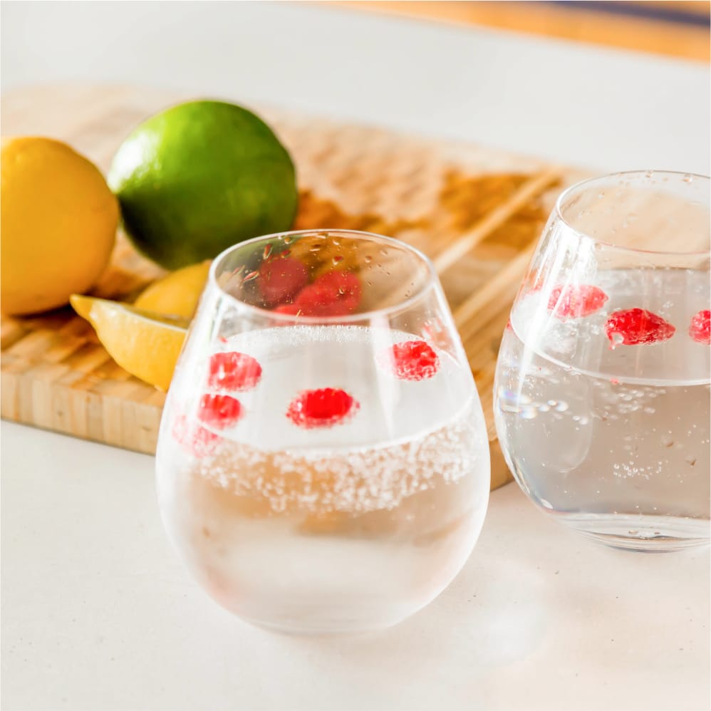 Delicious homemade drinks created using the SeltzaTap sit on a white kitchen counter. Two glasses with carbonated water and raspberries. Behind the glasses is a chopping board with a lemon and lime ready to be cut. Some slices of lemon are nearby.