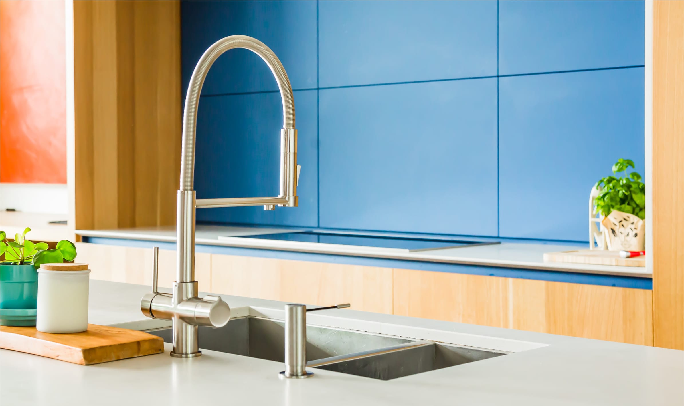 The Setzertap by Seltza, in a designer kitchen. The kitchen is blue and wood effect panelling. The tap is a brushed steel effect matching a steel sink. There are pot plants on the counter. Seltzatap brings carbonated water to your regular tap.