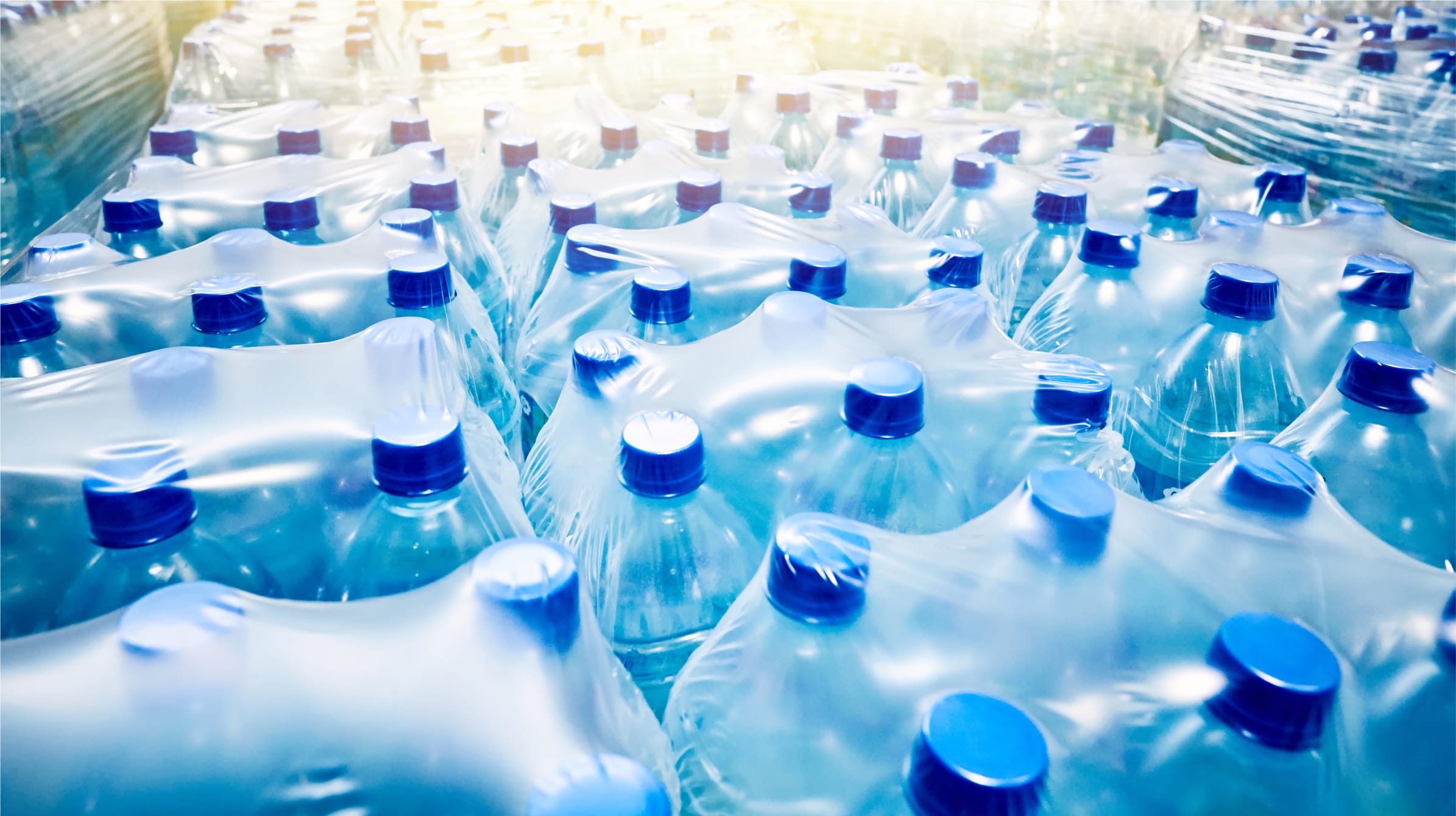 Packaged water waiting for delivery. The bottles are blue with darker blue caps, and go off into the distance becoming more blurred as they get further away. The bottled are polywrapped in packs of six waiting to be delivered.