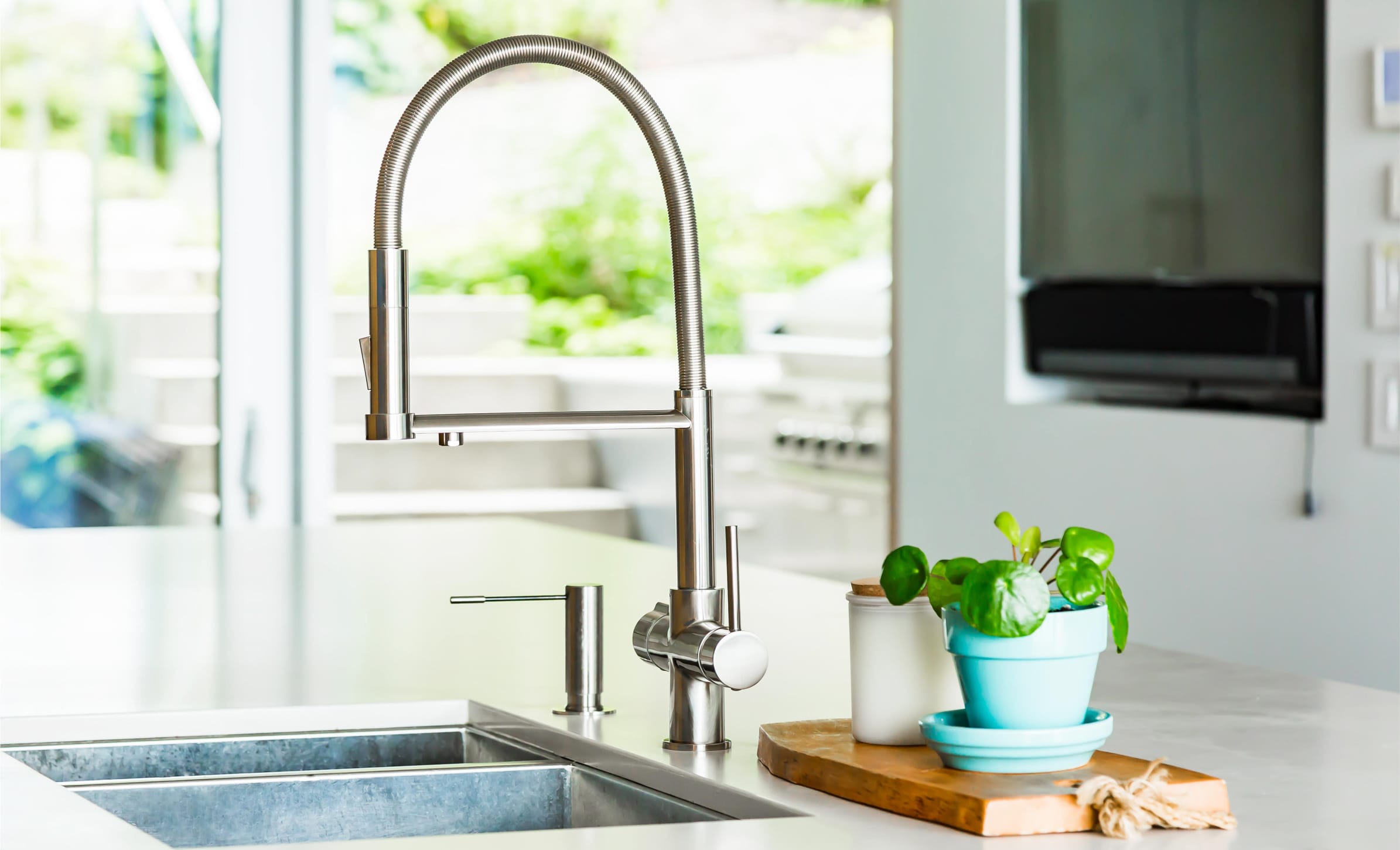 A brushed chrome Seltzatap installed an a brightly lit designer home. The countertop is white stone with some plant pots by the steel sink. In the background, a beautiful garden is visible through the patio windows.