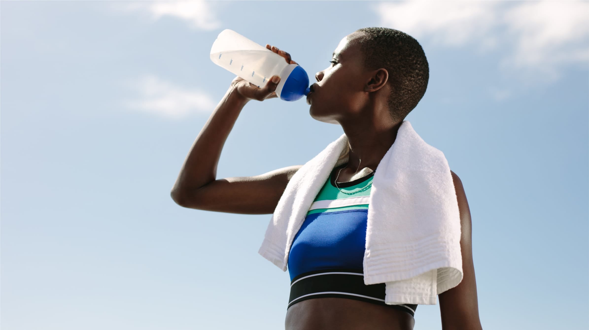 A woman in sports clothing stops during her workout to drink carbonated water from her water bottle. She has a towel around her neck and a bright blue sky is behind her.